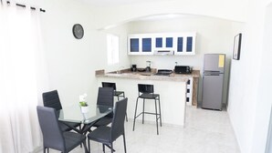 Een kleine keuken met witte kastjes, een koelkast, en een eethoek met vier stoelen en een glazen tafel.