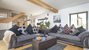 Ein modernes Wohnzimmer mit einer grauen Sofagarnitur, gemusterten Kissen, einem dunklen Hocker und einem großen Fenster mit Tageslicht.