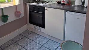 Una cocina con estufa a gas, horno y un refrigerador pequeño. Se ve una ventana con vistas al exterior, y también hay un cubo de basura.