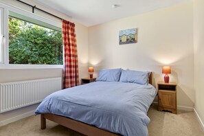 A bedroom with a bed, two bedside tables, a window with curtains, a radiator, and a wall painting. - A perfect for a relaxing getaway with family or friends (Tenby, Pembrokeshire)