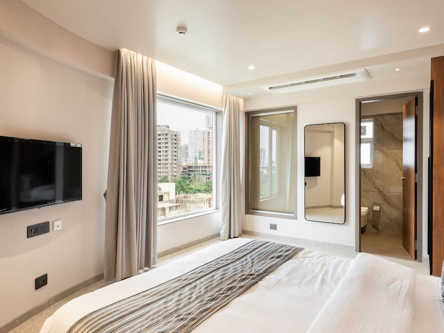A modern hotel room with a large bed, a flat-screen TV mounted on the wall, a large window with curtains, a bathroom visible through an open door, and a city view.