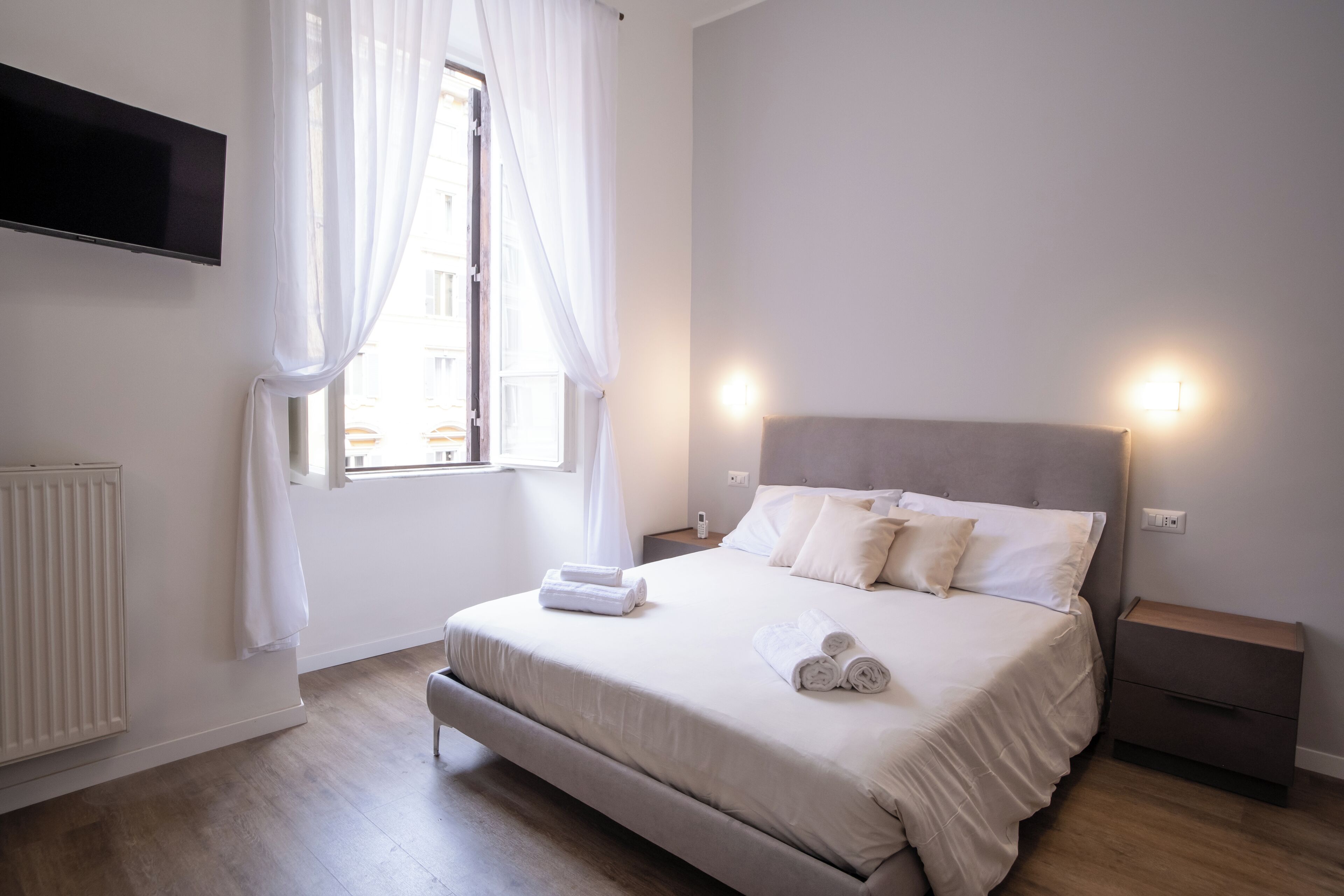 Ein Schlafzimmer mit einem Bett, einem Nachttisch, einem an der Wand befestigten Fernseher und einem Fenster mit Vorhängen.