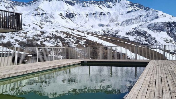 A wooden deck with a pool, surrounded by snow-covered mountains and a metal fence.