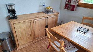 A small kitchen with wooden cabinets, a coffee maker, a trash can, and a dining table with chairs.