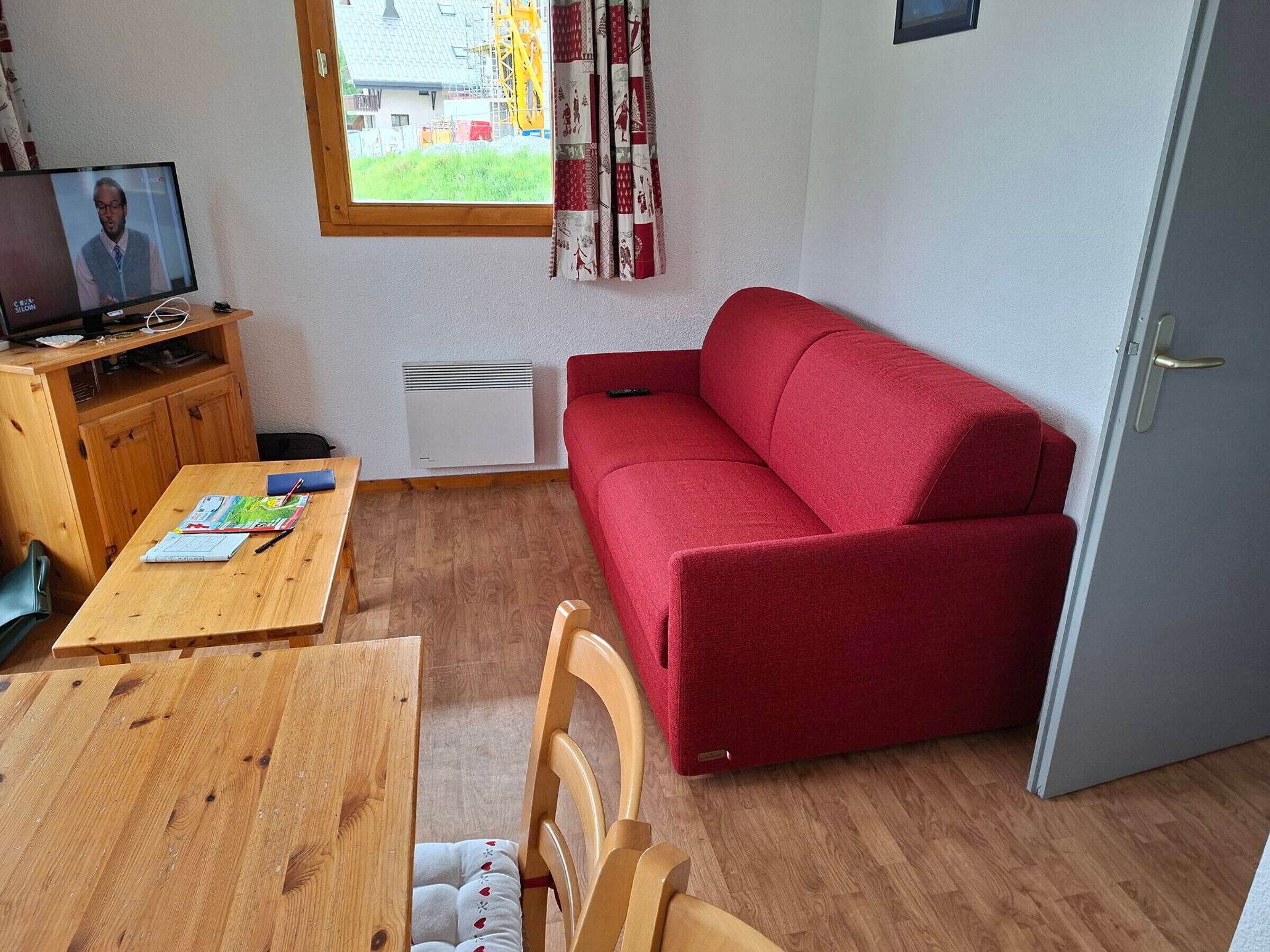 Un espacio habitable compacto con un sofá rojo, muebles de madera, un televisor sobre un soporte y una ventana con vistas a edificios y vegetación.