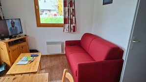 A compact living space with a red sofa, wooden furniture, a TV on a stand, and a window with a view of buildings and greenery.