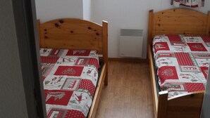 A small, cozy room with two wooden beds, each with a quilted bedspread. There is a window above the beds, and a wall-mounted heater between them.