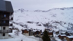 A snow-covered mountain village with multi-story buildings, including a hotel with balconies and a ski resort in the background.