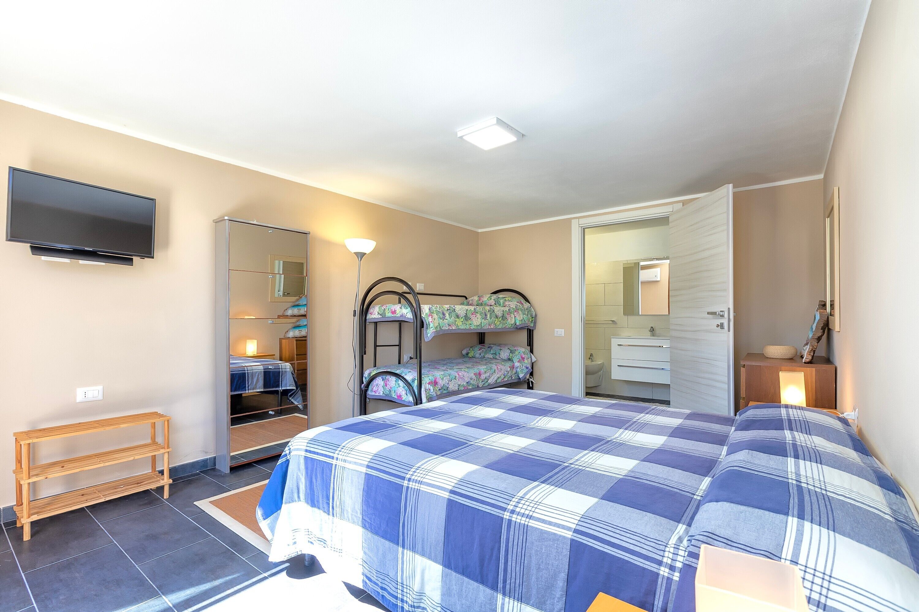 Un dormitorio con cama, televisión, espejo y baño visible a través de una puerta abierta.