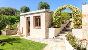 A modern building with a stone facade, a small entrance, and a garden with a statue and flowers. - Holiday Home 'Flora Sea View' with Sea View, Wi-Fi and Air Conditioning (Punta de su Torrione)
