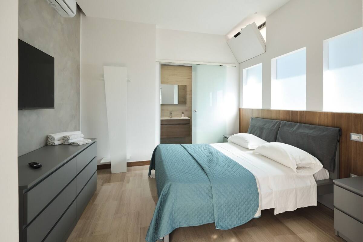 Quarto moderno com cama, mesas de cabeceira, televisão e banheiro visível por uma porta aberta.