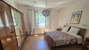 A bedroom with a bed, a window with curtains, a radiator, a wooden floor, a hanging light fixture, and a framed picture on the wall. - Air-conditioned villa with large garden, pets allowed (Palau-del-Vidre)