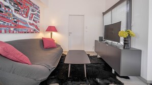 Ein modernes Wohnzimmer mit einem grauen Sofa, einem kleinen Tisch, einem Fernseher auf einer Kommode, einem Bild an der Wand und einer Tür im Hintergrund.