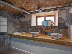 A bathroom with stone walls, a wooden vanity, two sinks, a mirror, and a window.