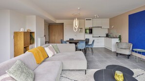Een moderne woonkamer met een grijze hoekbank, een eethoek met een ronde tafel en stoelen, en een keuken op de achtergrond.