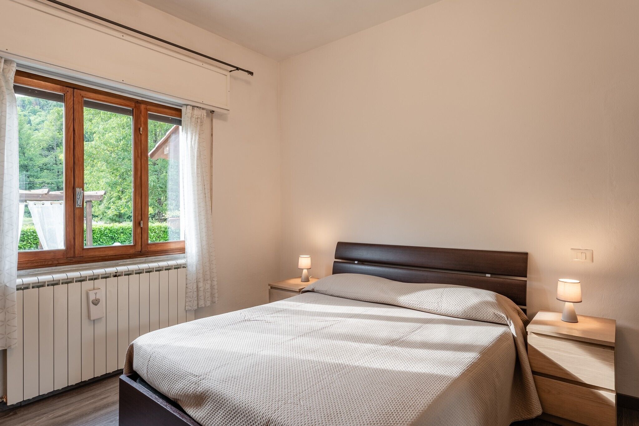 Un dormitorio con cama, dos lámparas de noche, una ventana con cortinas y un radiador.