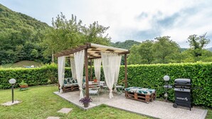 Een tuin met een pergola, terras, barbecue en een haag.