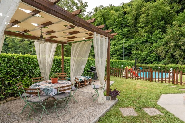A covered outdoor seating area with a table and chairs, a pergola with curtains, a pool, and a wooden fence.