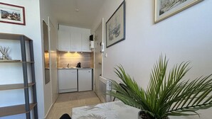 Cocina compacta con muebles blancos, una cafetera y un refrigerador pequeño. Hay una planta en maceta sobre la mesa y una escalera apoyada en la pared.