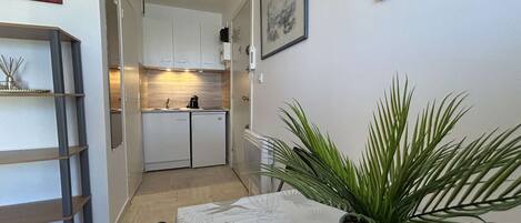 Une cuisine compacte avec des meubles blancs, une machine à café et un petit réfrigérateur. Il y a une plante en pot sur la table et une échelle appuyée contre le mur.