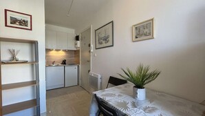 Una cocina compacta con armarios blancos, un refrigerador pequeño y una cafetera. Hay una mesa de comedor con mantel blanco y una planta en maceta.