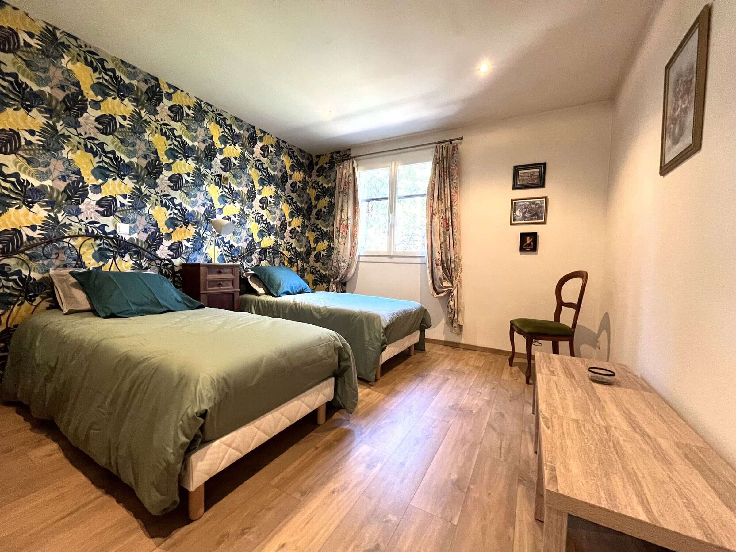 Une pièce avec deux lits, une table en bois, une chaise, une fenêtre avec des rideaux et un mur orné de tableaux.