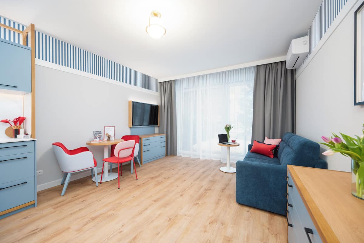 Ein modernes Wohnzimmer mit einem blauen Sofa, einem Essbereich mit roten und weißen Stühlen, einem Fernseher und einem kleinen Tisch mit einem Laptop.