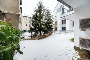 Un patio cubierto de nieve con un alto árbol de hoja perenne, una cerca de metal y un edificio blanco con una ventana y unidad de aire acondicionado.
