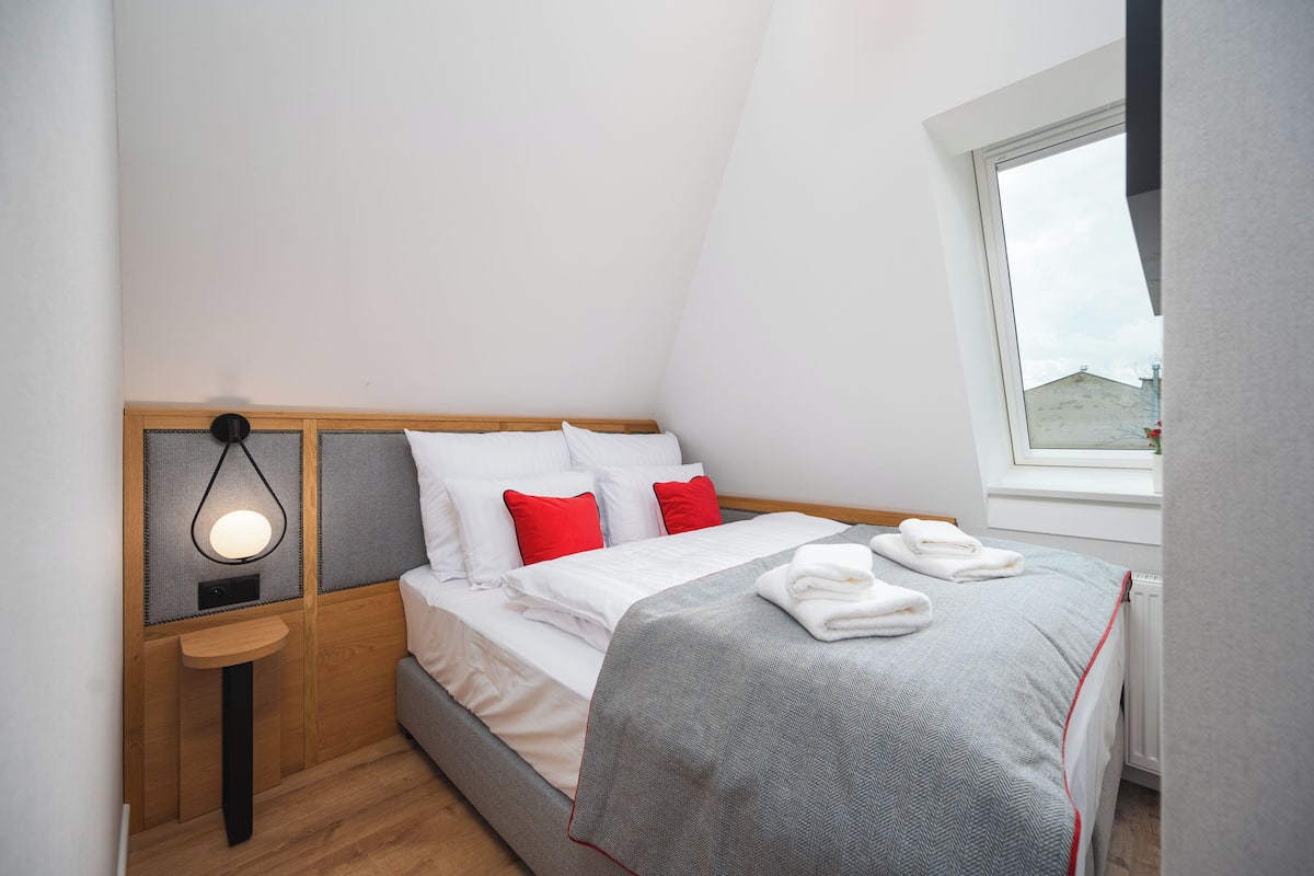 Ein ordentlich gemachtes Bett mit weißer und grauer Bettwäsche, roten Kissen und einer grauen Decke. Ein kleiner Nachttisch mit einer Lampe und ein Fenster mit Blick ins Freie sind ebenfalls zu sehen.
