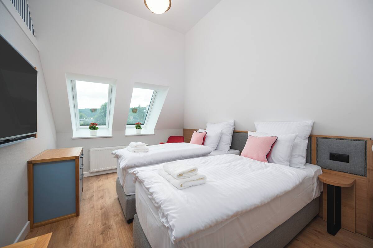 Ein modernes Hotelzimmer mit zwei Betten, einem kleinen Tisch, einem Fernseher und einem Fenster mit Blick auf Grünflächen.