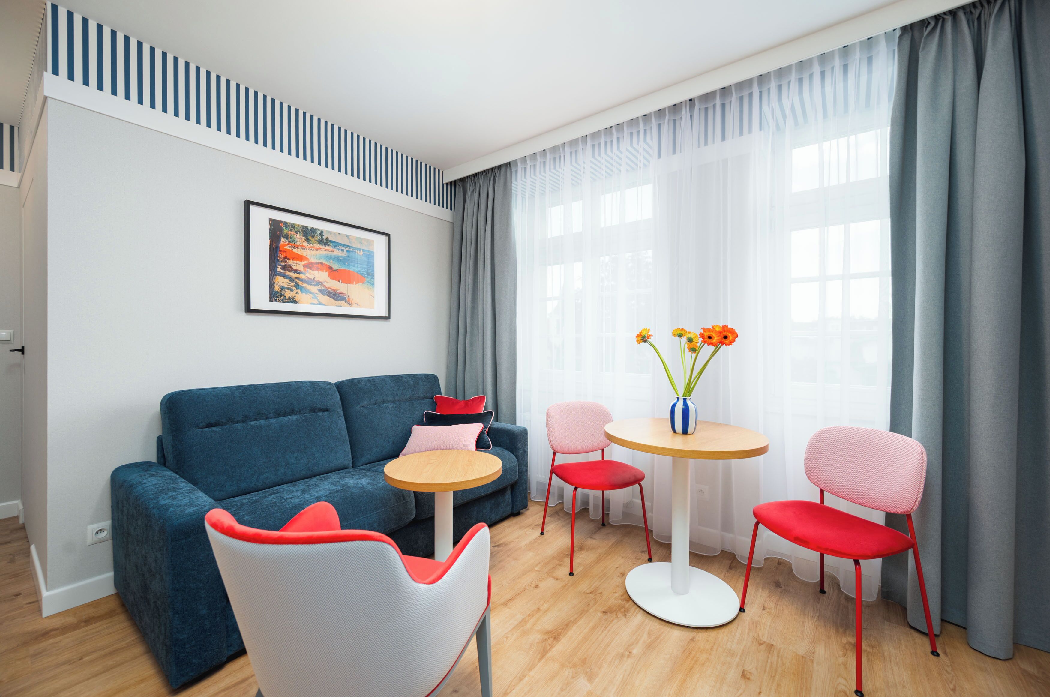 Ein modernes Wohnzimmer mit einem blauen Sofa, roten und weißen Stühlen, einem kleinen runden Tisch und einem Bild an der Wand.