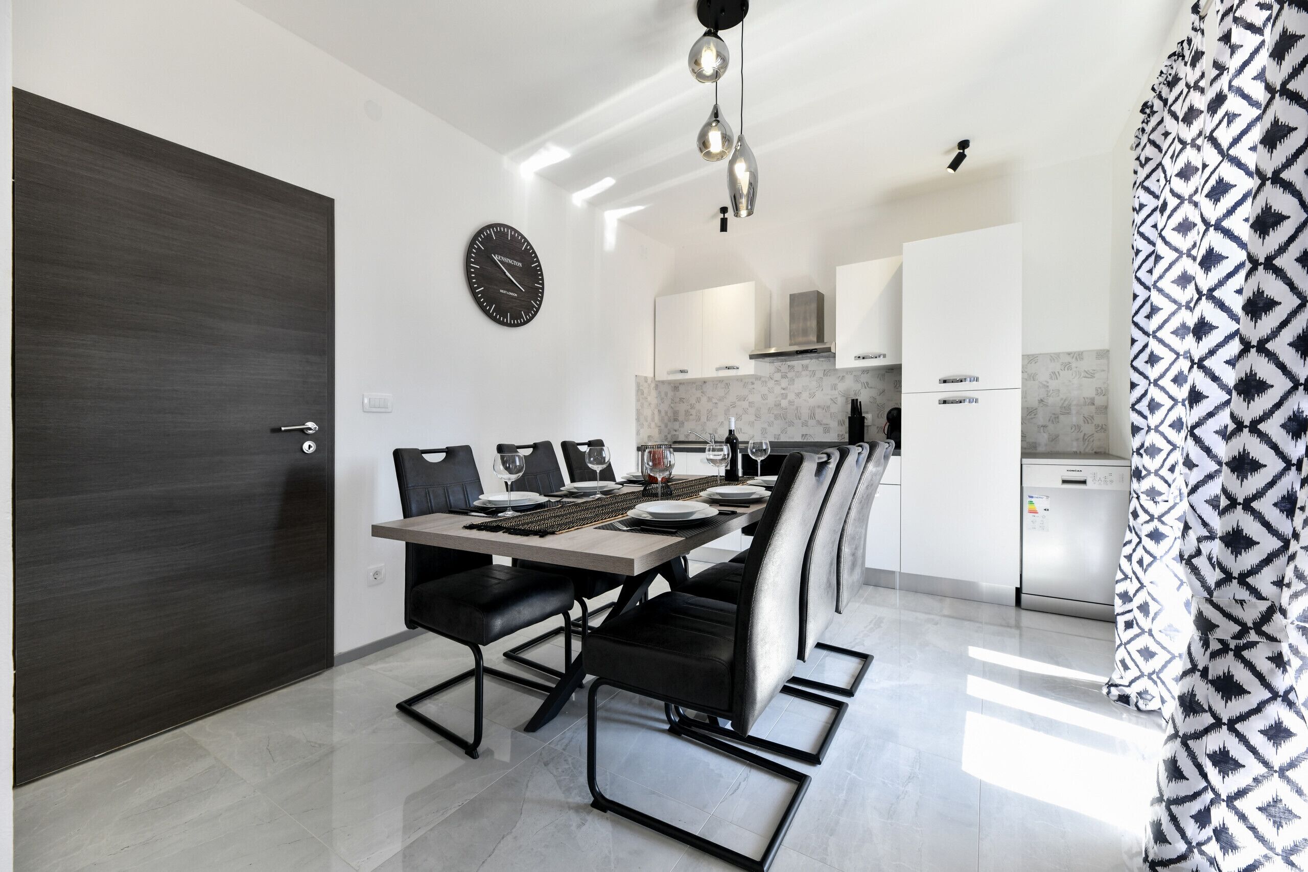 Cucina moderna con mobili bianchi, una porta in legno scuro e un'area pranzo con sedie nere e un tavolo imbandito per quattro.