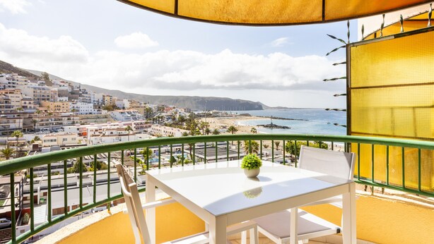 A balcony with a white table and chairs, a potted plant, and a view of a coastal town and sea. - Stunning sea view apt in Los Cristianos (Los Cristianos)