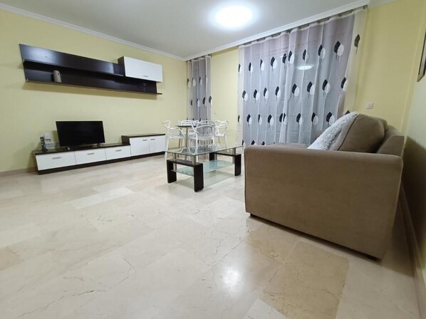 Una sala de estar con un sofá color beige, una mesa de centro de vidrio, un televisor sobre un mueble bajo y una zona de comedor con sillas y una mesa.
