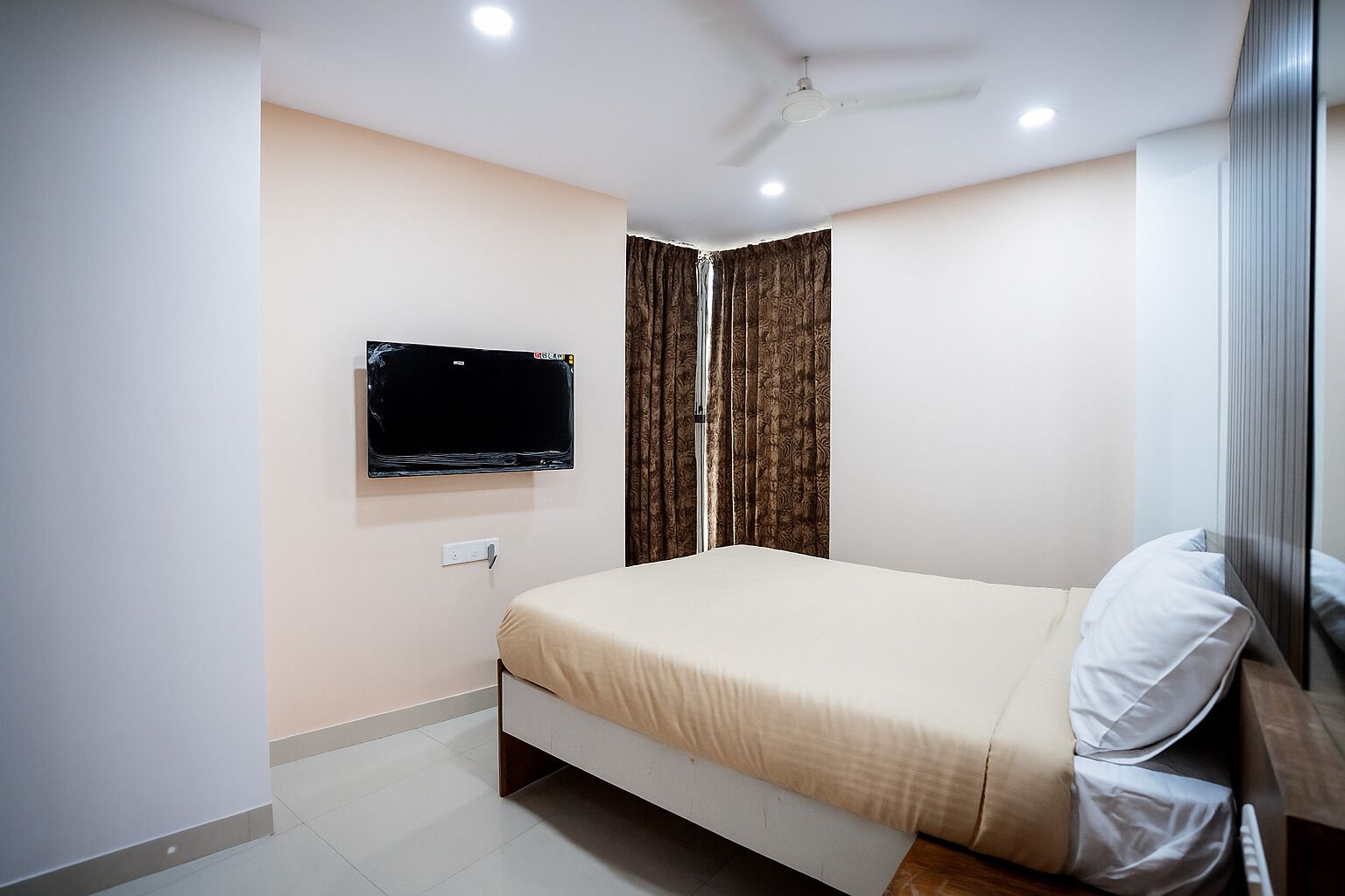 A bedroom with a bed, a TV mounted on the wall, a ceiling fan, and curtains.