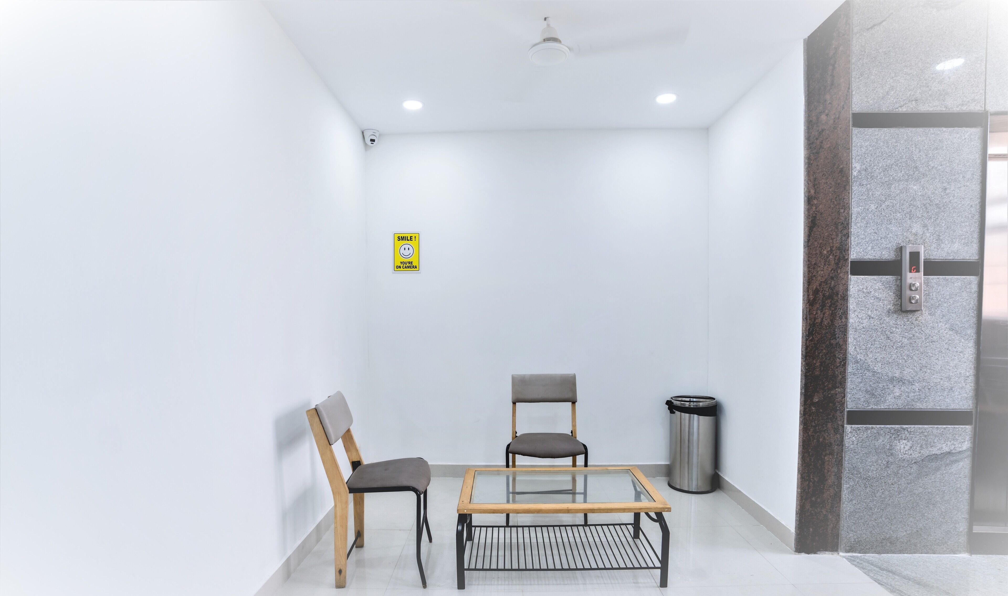 Un área de espera moderna con una mesa de vidrio, dos sillas, un cesto de basura y una cámara de seguridad.