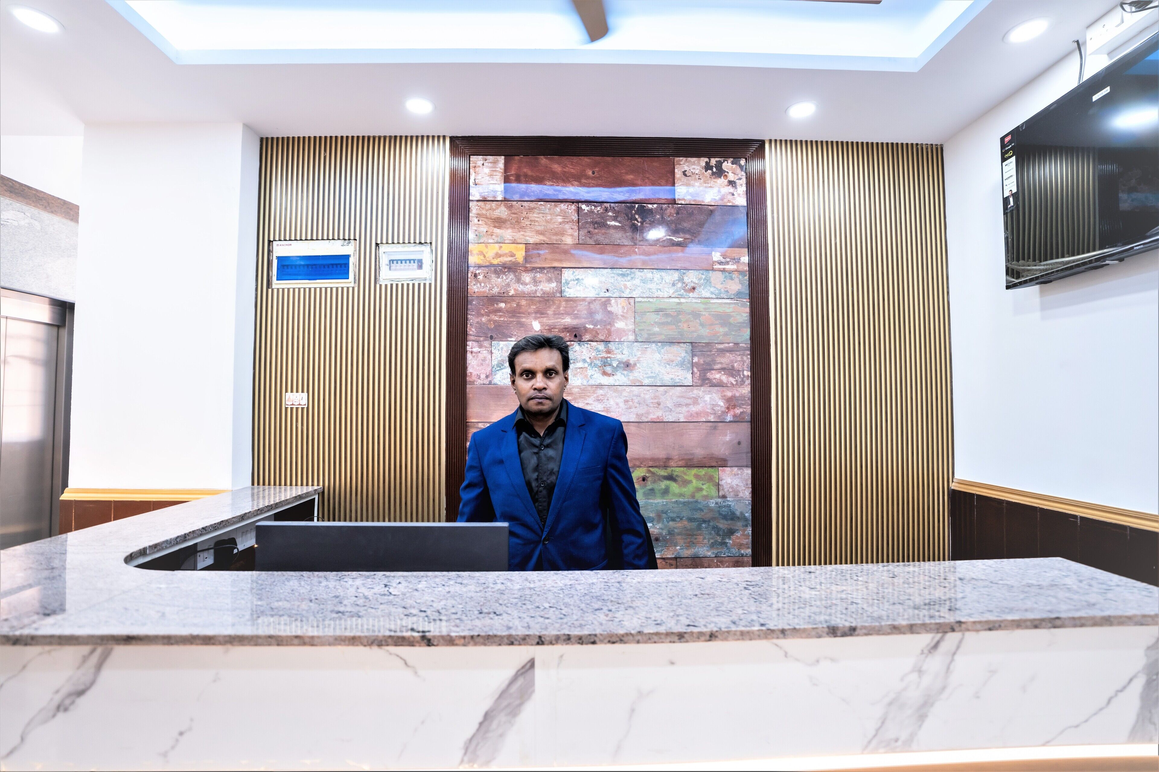 En man i en blå kostym står vid receptionsdisken i en modern entréhall med en vägg av sten och träpanel.