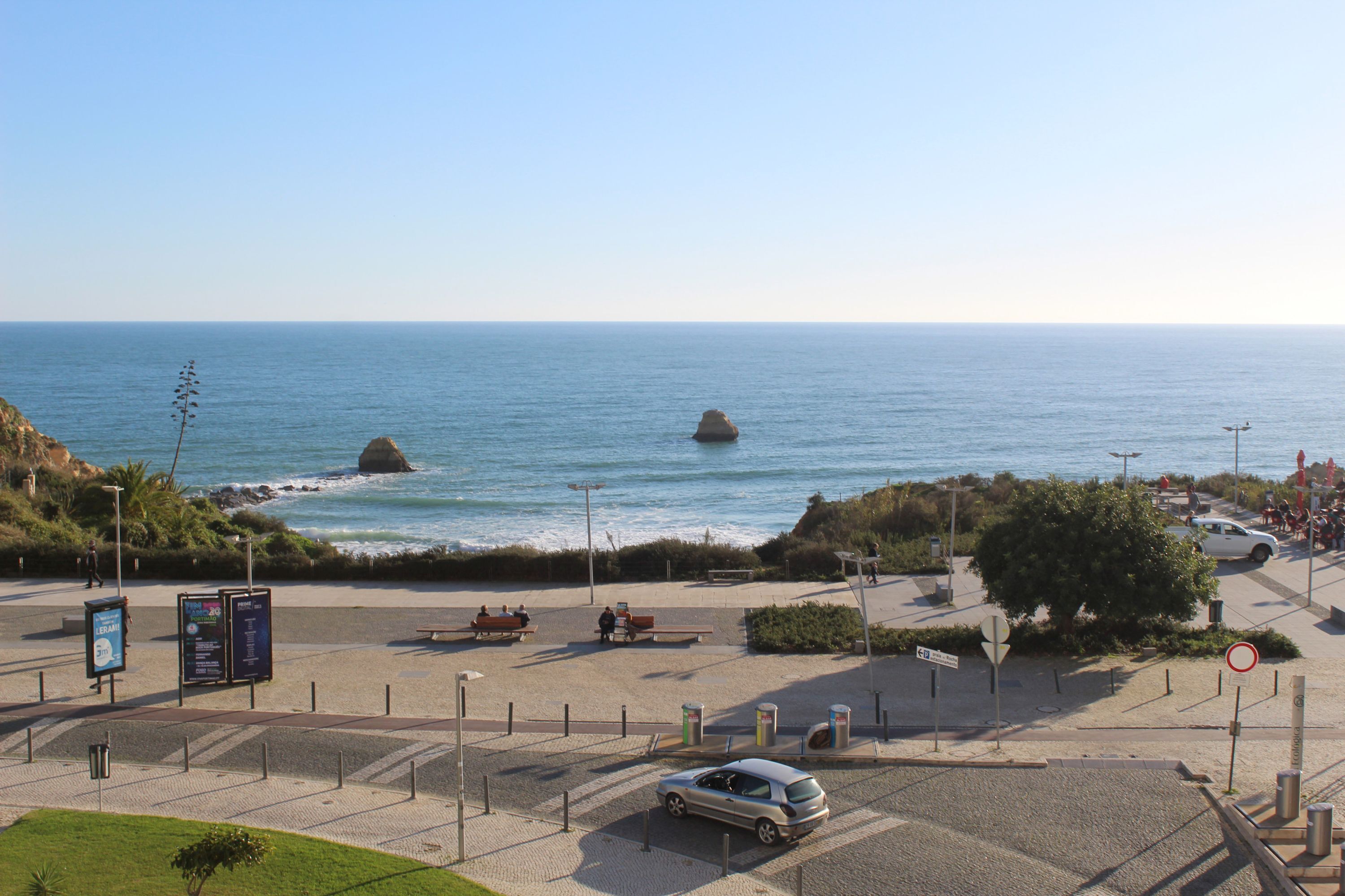Eine Küstenstraße mit geparktem Auto, einem Fußweg und Blick auf das Meer mit Felsen.