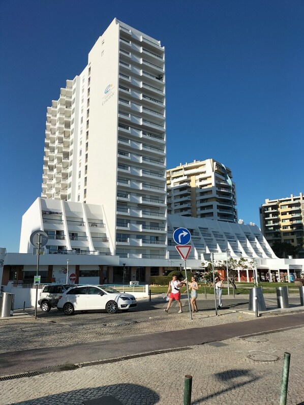 Un hôtel moderne, composé d’un immeuble de grande hauteur doté de nombreux balcons, d’une zone piétonne avec une signalisation directionnelle et d’un parking avec des voitures.