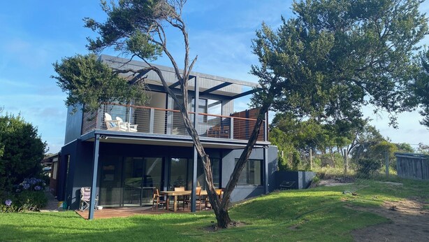 Ein modernes, zweistöckiges Haus mit einer überdachten Terrasse, einem Essbereich und einem großen Baum im Vorgarten.