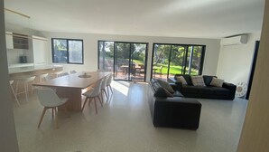 Ein modernes Wohnzimmer mit einem großen Esstisch, einem schwarzen Sofa und Blick auf einen Garten durch große Fenster.