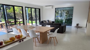 Ein modernes Wohnzimmer mit einem Holzespeisetisch, weißen Stühlen, einem dunkelgrauen Sofa und einem großen Fenster mit Blick auf einen Garten.
