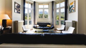A modern living room with a black sofa, white chairs, and a glass coffee table. Large windows offer a view of a mountainous landscape. - Superb apartment in Mexican house (Barcelonnette)