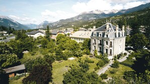 A large, historic building with a prominent tower, surrounded by lush greenery and mountains in the background. - Superb apartment in Mexican house (Barcelonnette)