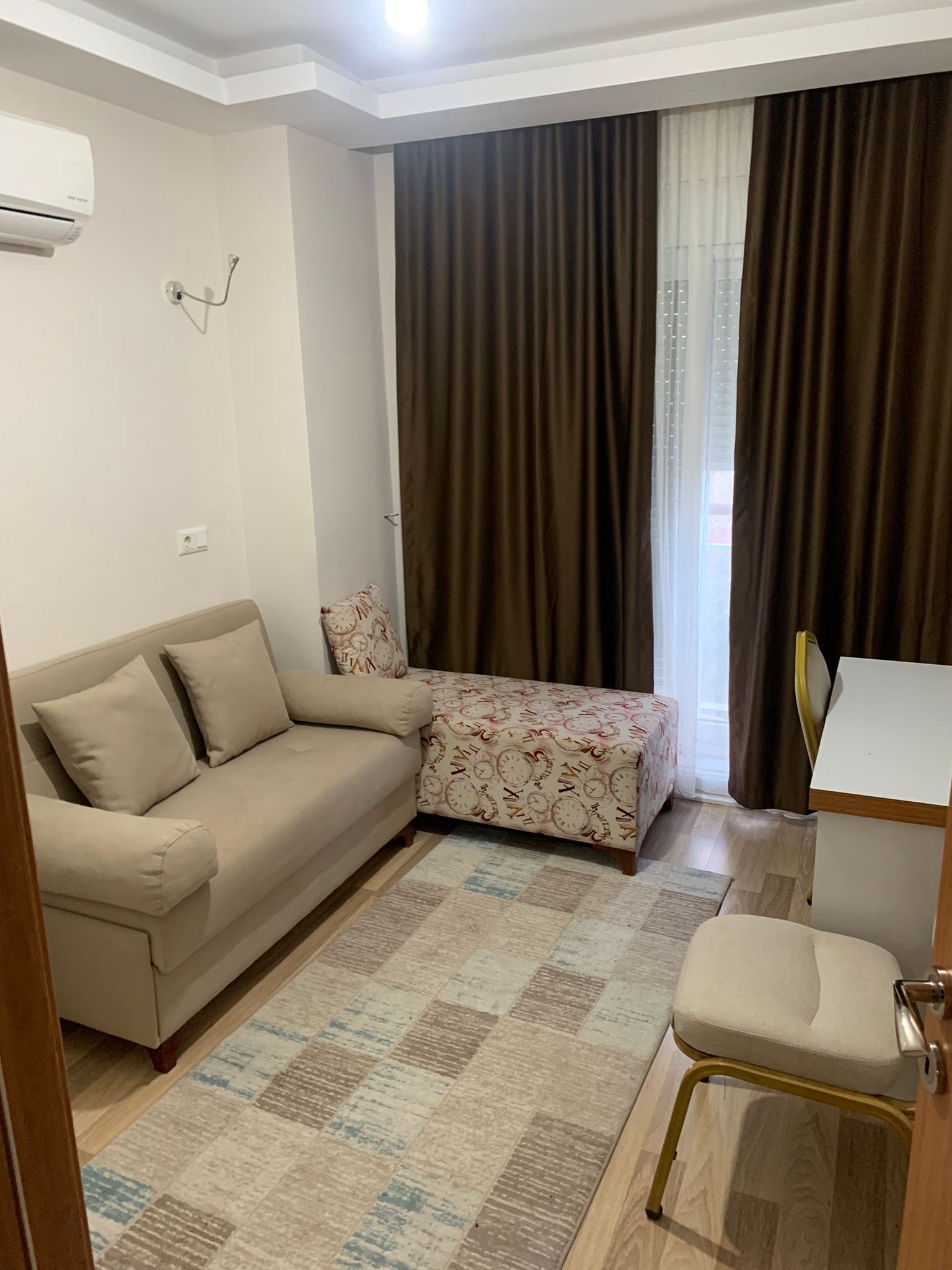 A small, well-lit room with a beige sofa, a patterned ottoman, a small table, and a chair. There is a window with curtains and an air conditioning unit on the wall.