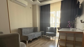 A modern living room with a grey sofa, a small round table, and a wooden coffee table. There is a built-in air conditioning unit on the wall and a window with curtains. - Nativo Hotel (Antalya)