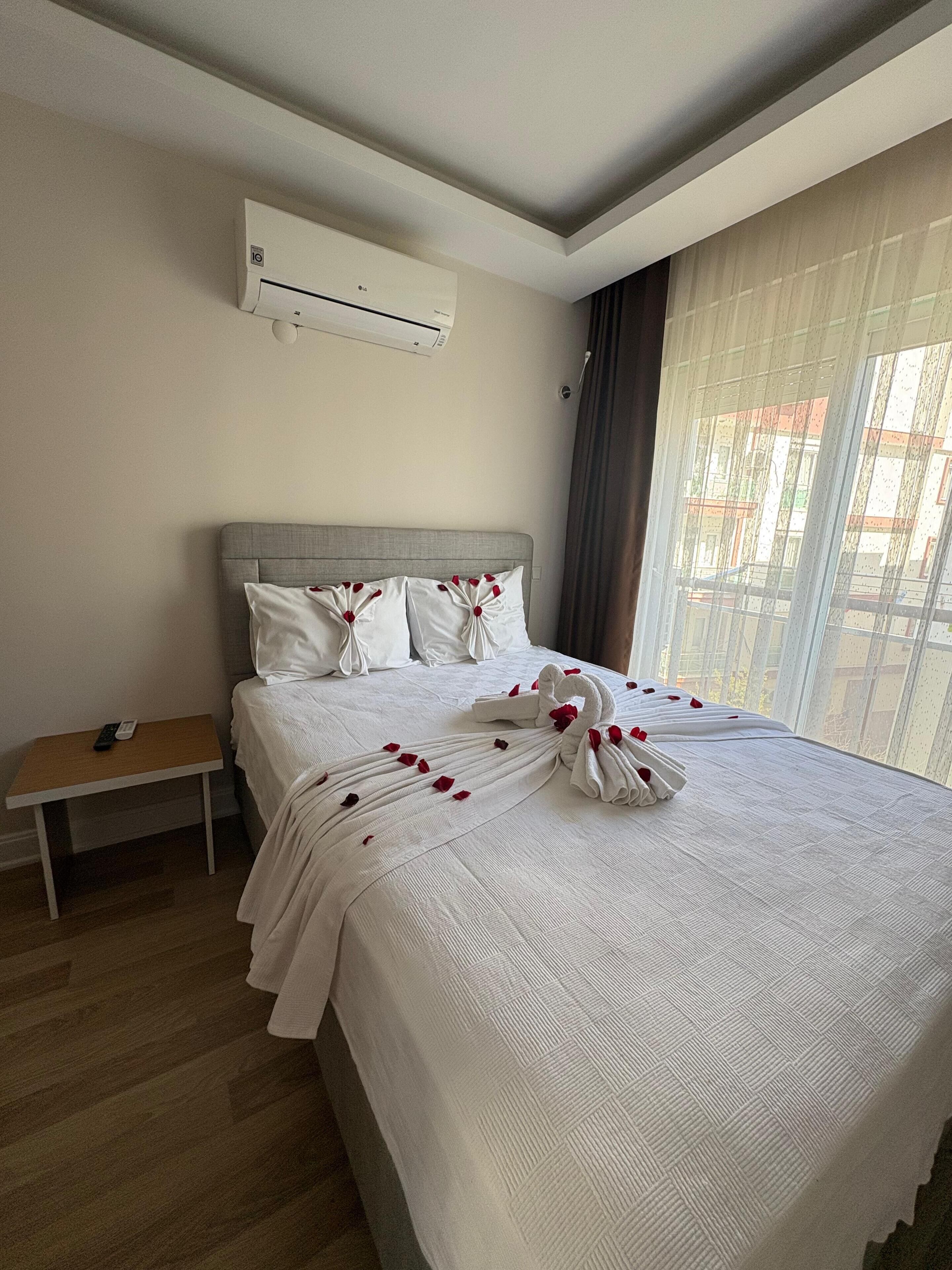 Una cama bien hecha con una disposición de toallas en forma de cisne y pétalos de rosa, una mesita de noche con un control remoto y un equipo de aire acondicionado en la pared.