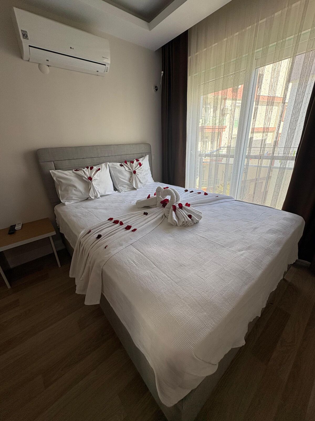 A hotel room with a bed, a bedside table, an air conditioning unit, and a window with curtains.