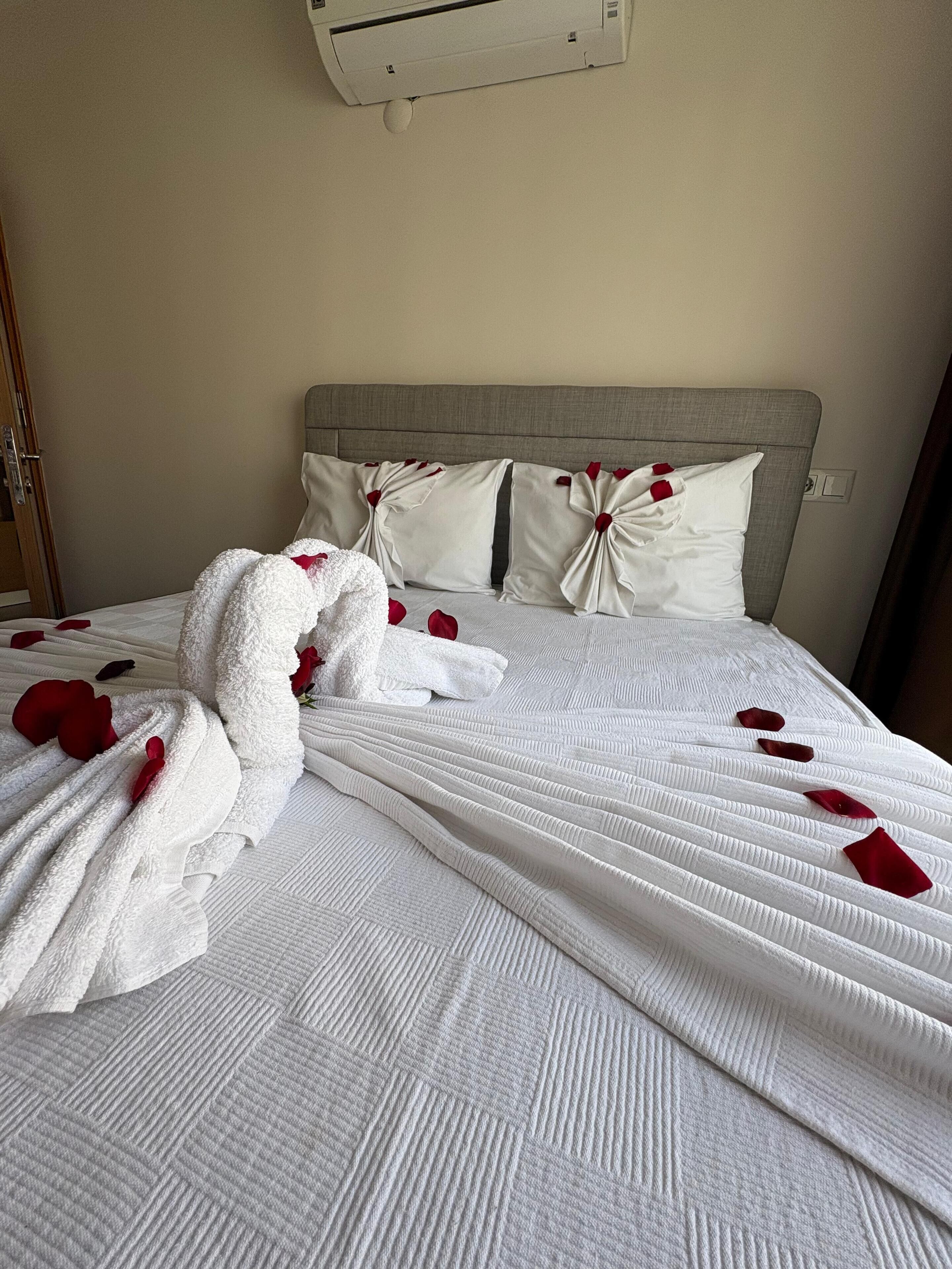 Una cama impecablemente arreglada con una colcha blanca y almohadas, una toalla blanca y pétalos de rosa roja esparcidos sobre la cama.