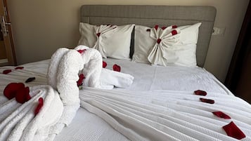 Una cama impecablemente arreglada con una colcha blanca y almohadas, una toalla blanca y pétalos de rosa roja esparcidos sobre la cama.